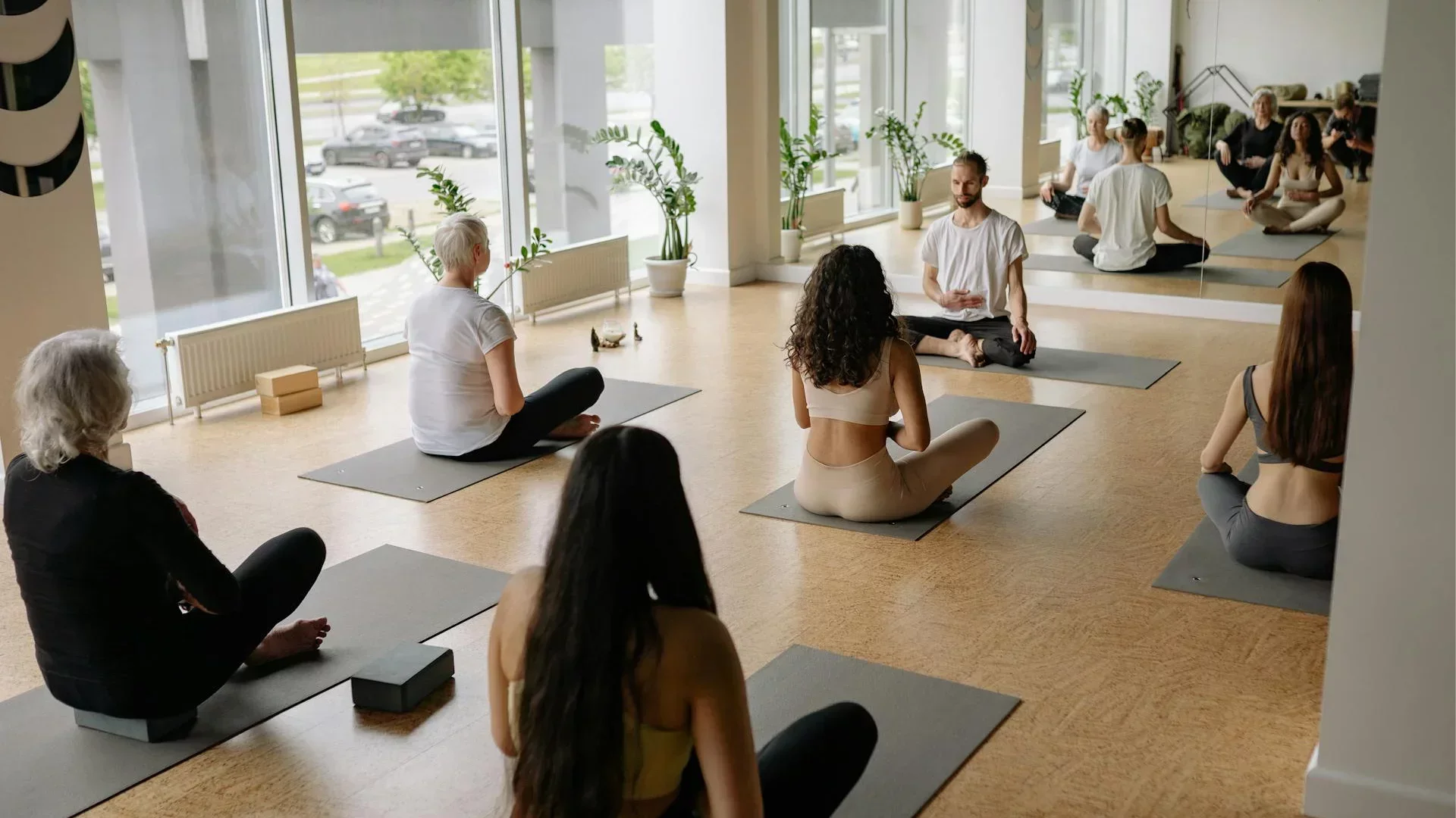 Yoga instructor leading a seated class with diverse students in a bright, modern studio with large windows and mirrors