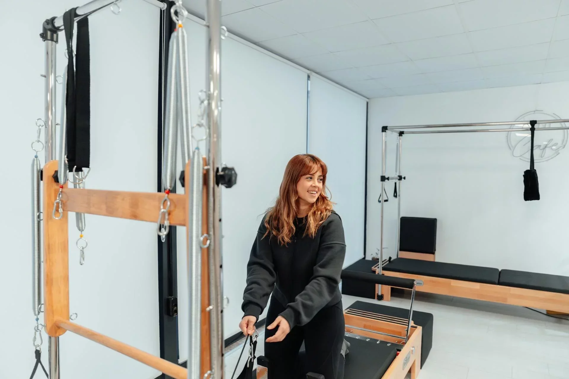Smiling Pilates instructor setting up reformer equipment in a bright modern Pilates studio with professional apparatus and minimalist design.