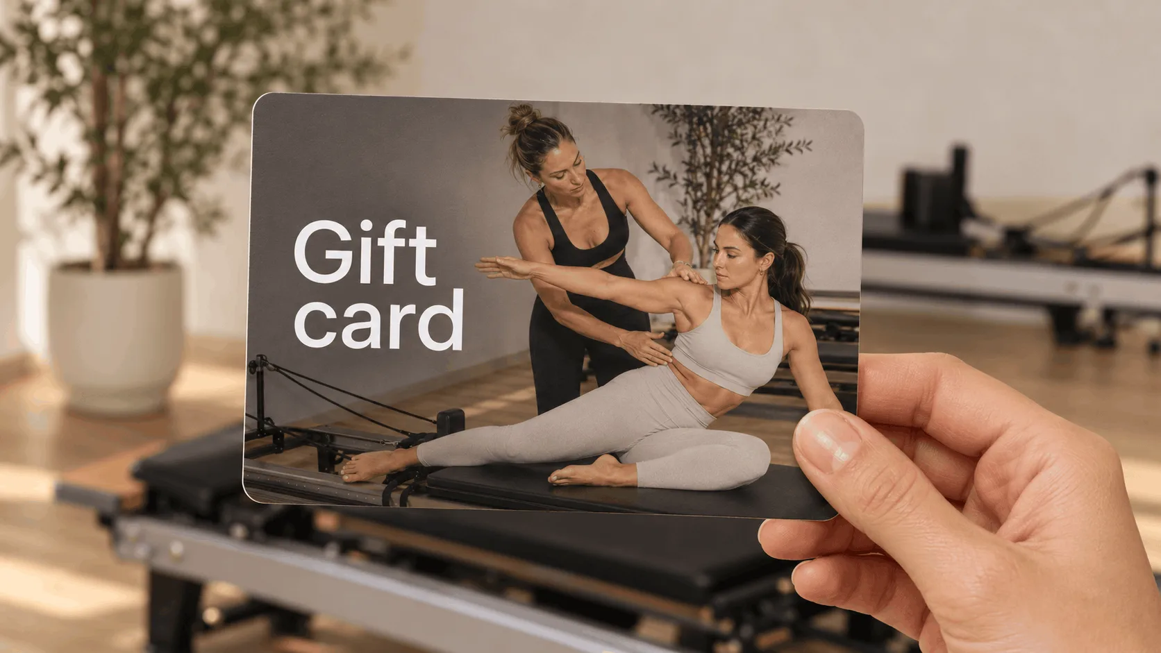 Fitness studio gift card held in front of Pilates reformers, showing an instructor guiding a client on a reformer.
