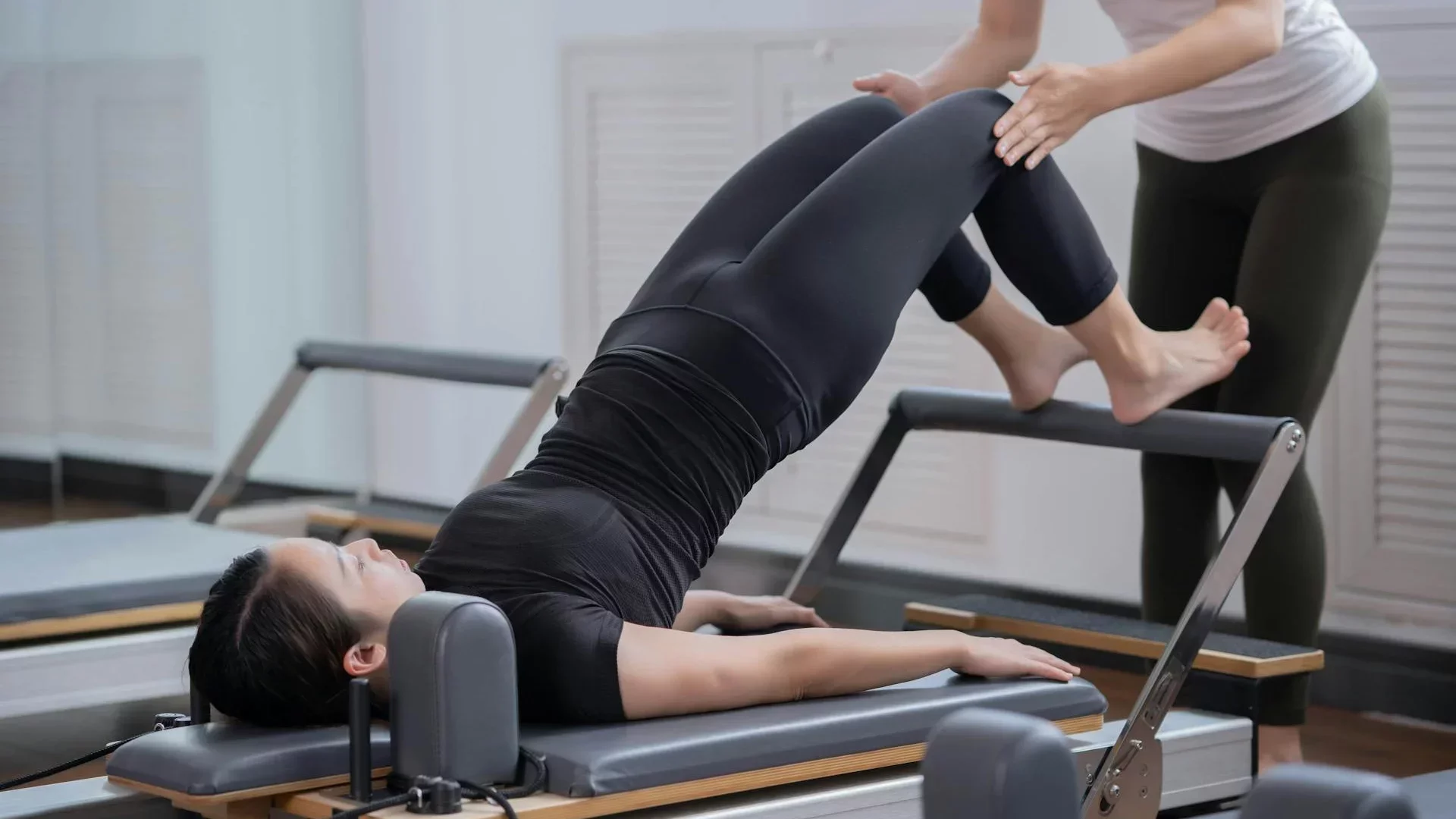 Pilates instructor assisting a client on a reformer machine during a bridge exercise in a modern studio setting.